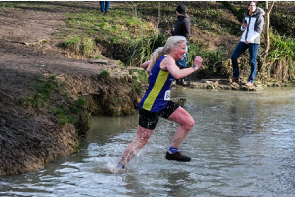 Cross country, Pittville Park cross country, Running, Runner, A female runner in a purple vest and black shorts runs through a stream with water splashing up her legs