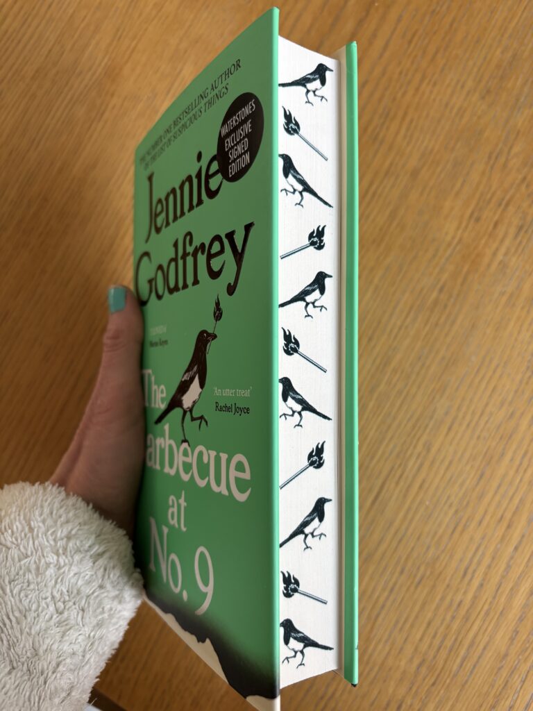 The Barbecue at No.9 by Jennie Godfrey, The Barbecue at No.9, Jennie Godfrey, Book review, A woman's hand holds a green book called The Barbecue at No.9 by Jennie Godfrey to show the edges of the pages which are decorated with illustrations of magpies and lit matches