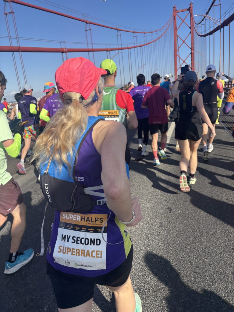Lisbon half marathon, Runners, Running, Pont de 25 Abril, Lisbon, Silent Sunday, My Sunday Photo, A crowd of runners run across a big red bridge against a blue sky with a female runner in a pink hat and a sign on her back saying My Second Superrace in the foreground