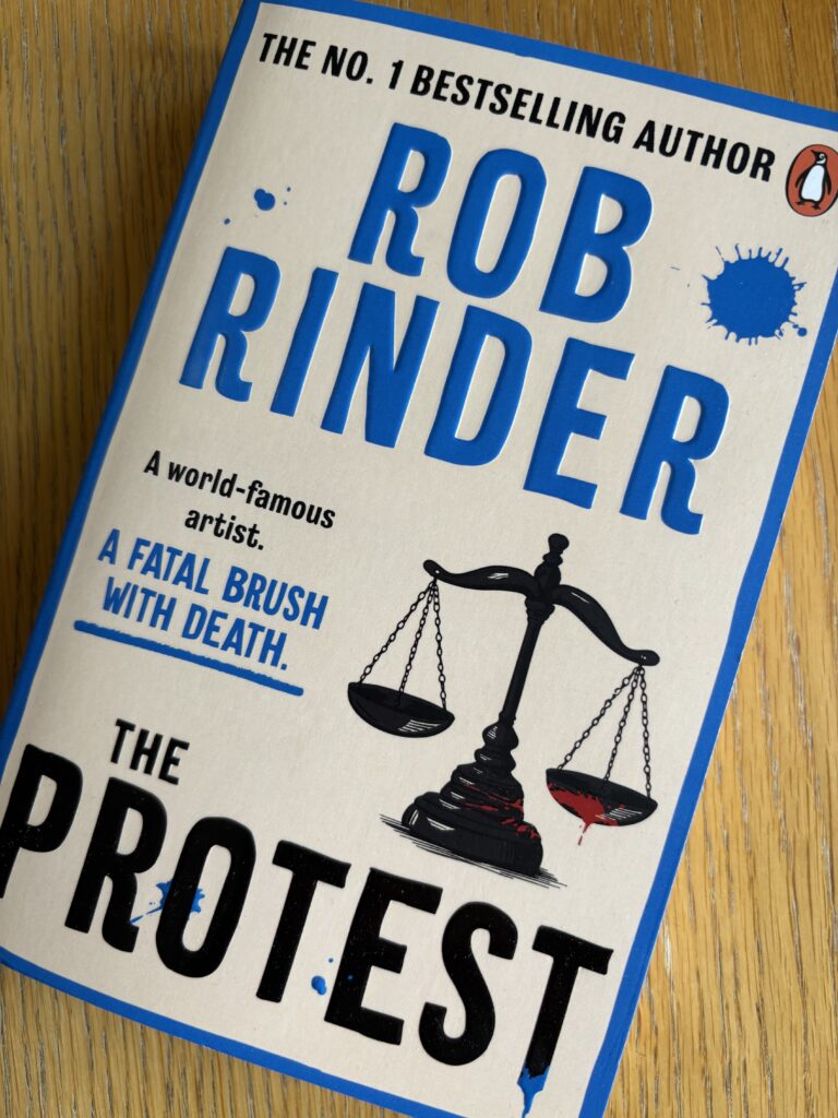 The Protest by Rob Rinder, The Protest, Rob Rinder, A light coloured book called The Protest by Rob Rinder with blue edges and a blob of blue paint and an illustration of the scales of justice on the front sits on a wooden table
