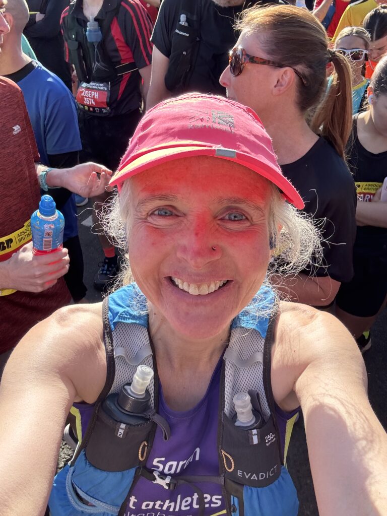 Newport marathon, Running, Runner, Marathon runner, Head and shoulders of a smiling female runner standing in the sun wearing a pink hat and a hydration vest with crowds of runners behind her