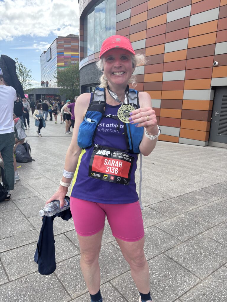 Newport marathon, Marathon medal, Runner, Running, A smiling female runner wearing pink shorts and a pink hat stands on the pavement outside a shopping centre holding the medal around her neck