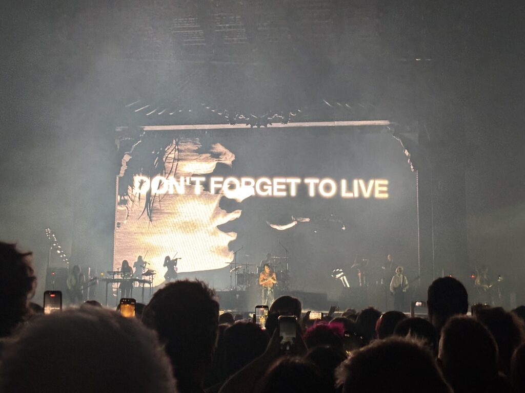 Yungblud, Yungblud live, Idols World Tour, Birmingham Utilita Arena, Birmingham, A shirtless man stands on a dark stage in front of a large crowd with his on a screen on the back with the words Don't forget to live