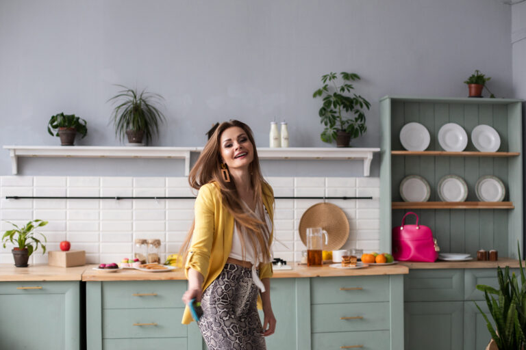 KItchen, Kitchen colours, AI image of a young woman with long brown hair in a spacious kitchen with grey-green cupboards and white tiles with various plants and white plates on the surfaces and shelving