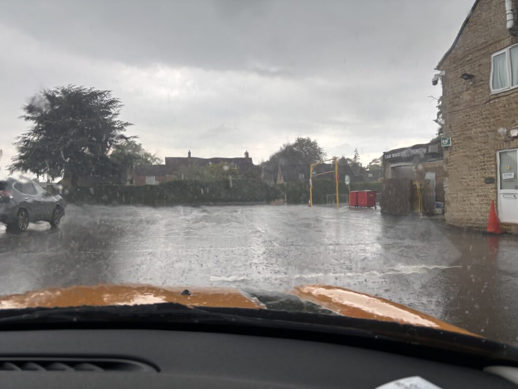 Bourton mile, Bourton-on-the-Water, Rainy car park, Orange and black bonnet of a car is visible through the windscreen looking out on a car park with torrential rain and a very dark cloudy sky
