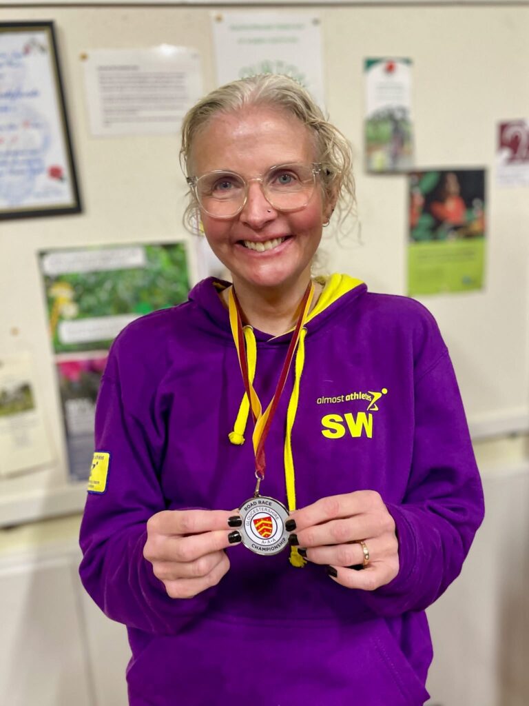 Bourton mile, Medal, A smiling woman in glasses wearing a purple hoodie wears a silver road race medal