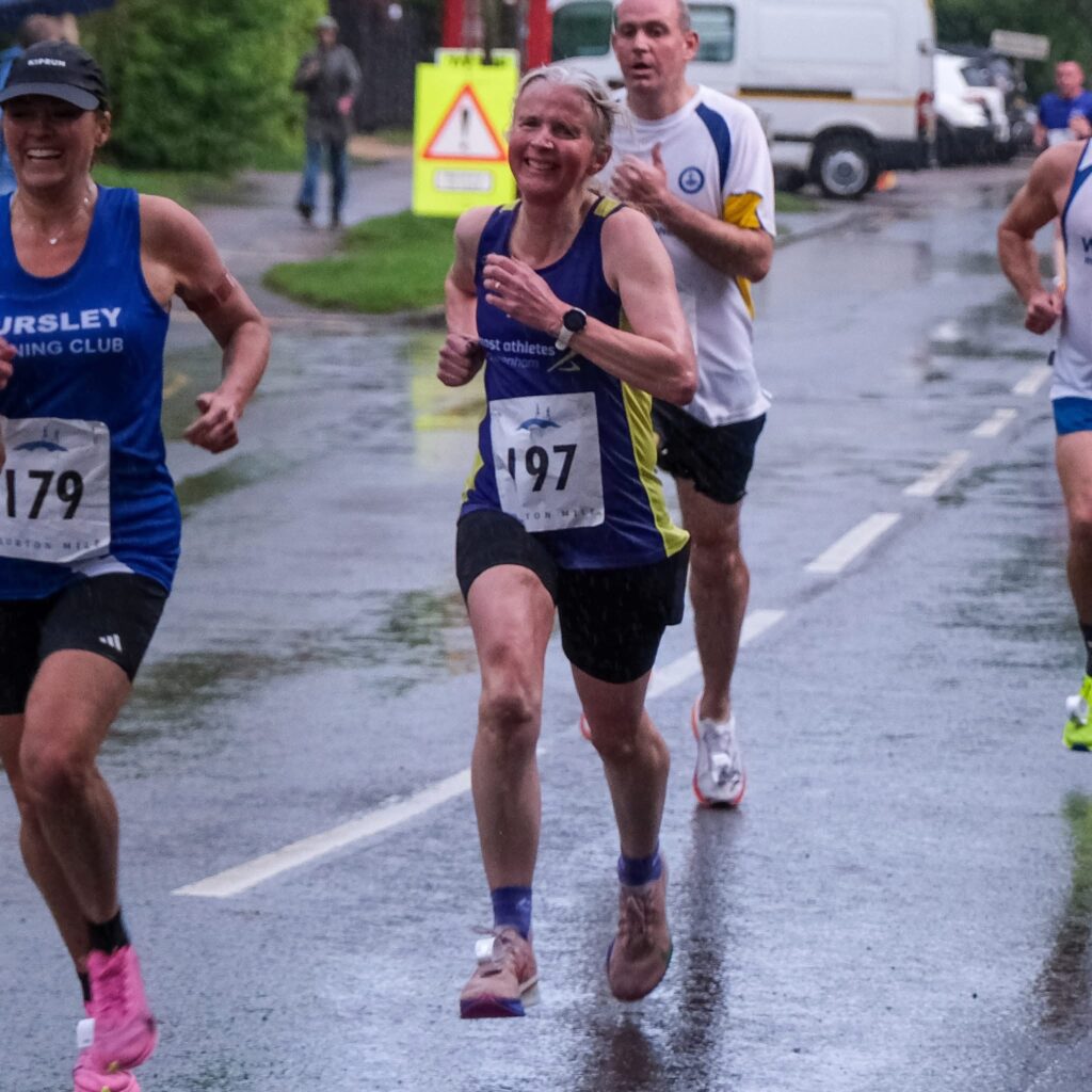 Bourton mile, Running, Runners, A grimacing female runner in a purple vest on a very wet road tries to catch a female runner in a blue vest with male runners in the background