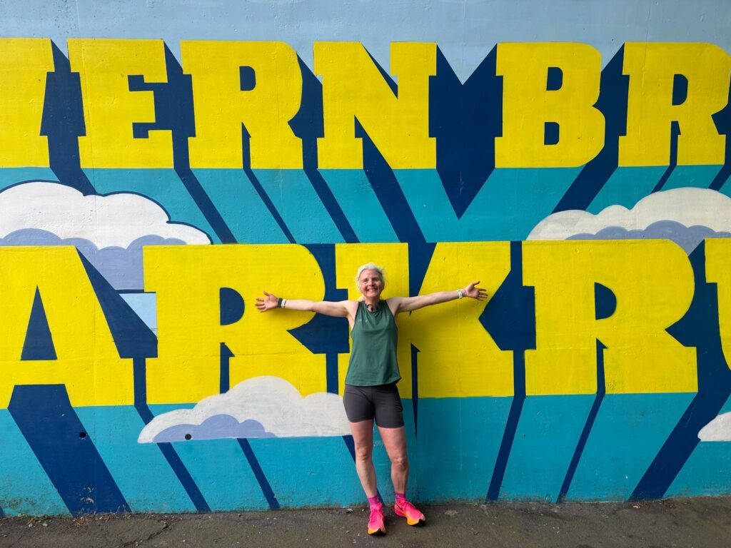 Severn Bridge parkrun, Parkrun, Parkrun tourism, Silent Sunday, My Sunday Photo, A woman in a green running vest and bright pink trainers stands with her arms spread against a wall with Severn Bridge Parkrun in big blue and yellow letters