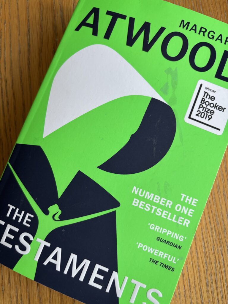 The Testaments by Margaret Atwood, The Testaments, Margaret Atwood, Book review, A green book called The Testaments by Margaret Atwood with a stylised image of a woman's head and shoulders in a bonnet and cape sits on a wooden table
