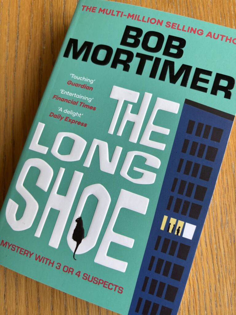 The Long Shoe by Bob Mortimer, The Long Shoe, Bob Mortimer, Bob Mortimer, A tourquoise book with an illustration of a towerblock with one floor lit up called The Long Shoe by Bob Mortimer with a cat sitting on the O of shoe sits on a wooden table