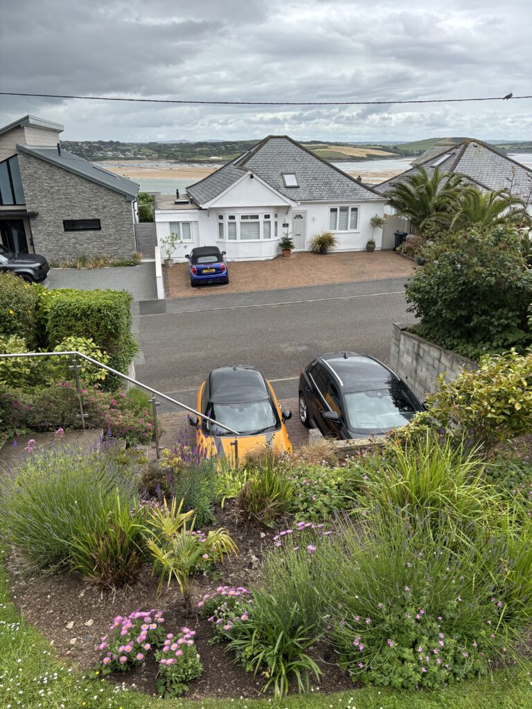 42 Dennis Road, Padstow, Cornwall, Holiday, A garden with various flowers in front of a partially visible black car and a partially visible orange car with a white house and a sandy estuary in the background
