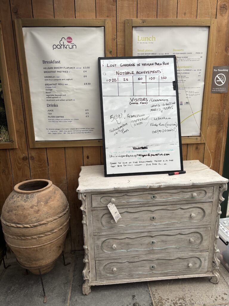 The Lost Gardens of Heligan parkrun, Parkrun tourism, A chest of drawers and a big terracotta pot with a whiteboard for people to write their achievements and a sign saying parkrun breakfast