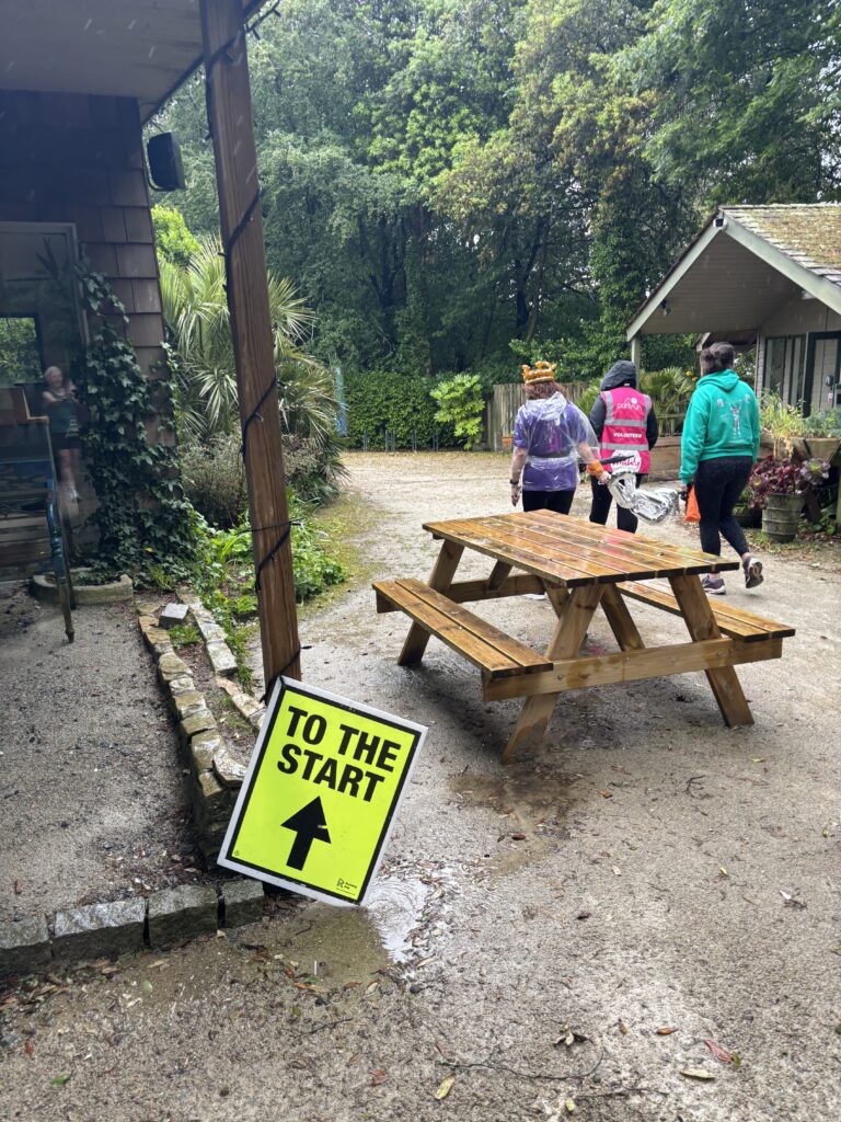 The Lost Gardens of Heligan parkrun, Parkrun, Parkrun tourism, Cornwall, Trees and a wet picnic table with muddy ground covered in puddles and three people walking away from the camera behind a yellow sign which says To the start with an arrow