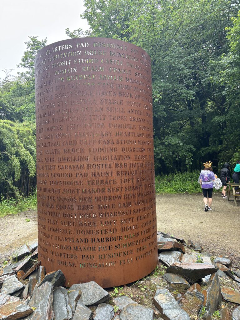 The Lost Gardens of Heligan parkrun, Parkrun tourism, Cornwall, A tall rusty cylinder surrounded by rocks withe words punched into it surrounded by trees and wet ground