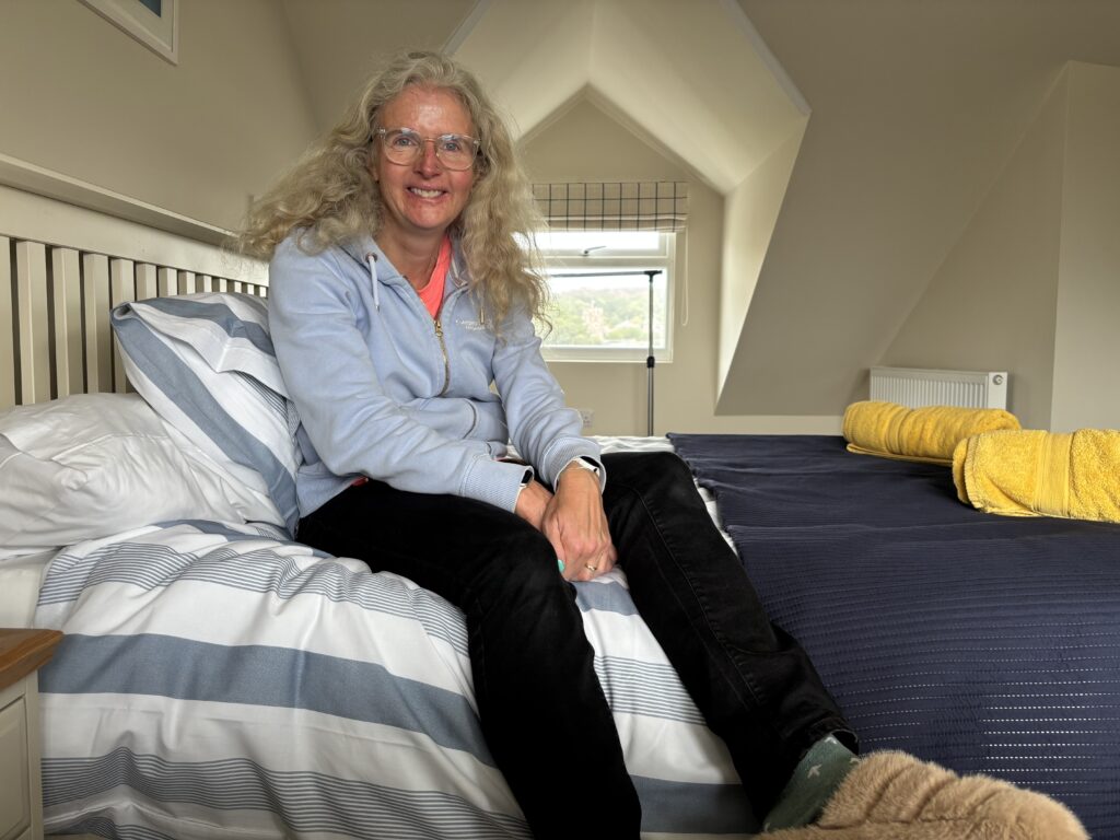 Holiday house, 8 New Street, Padstow, Cornwall, A smiling woman with blonde hair and glasses wearing a light blue hoodie sits on a bed wiht blue and white striped bedding with a window in the background