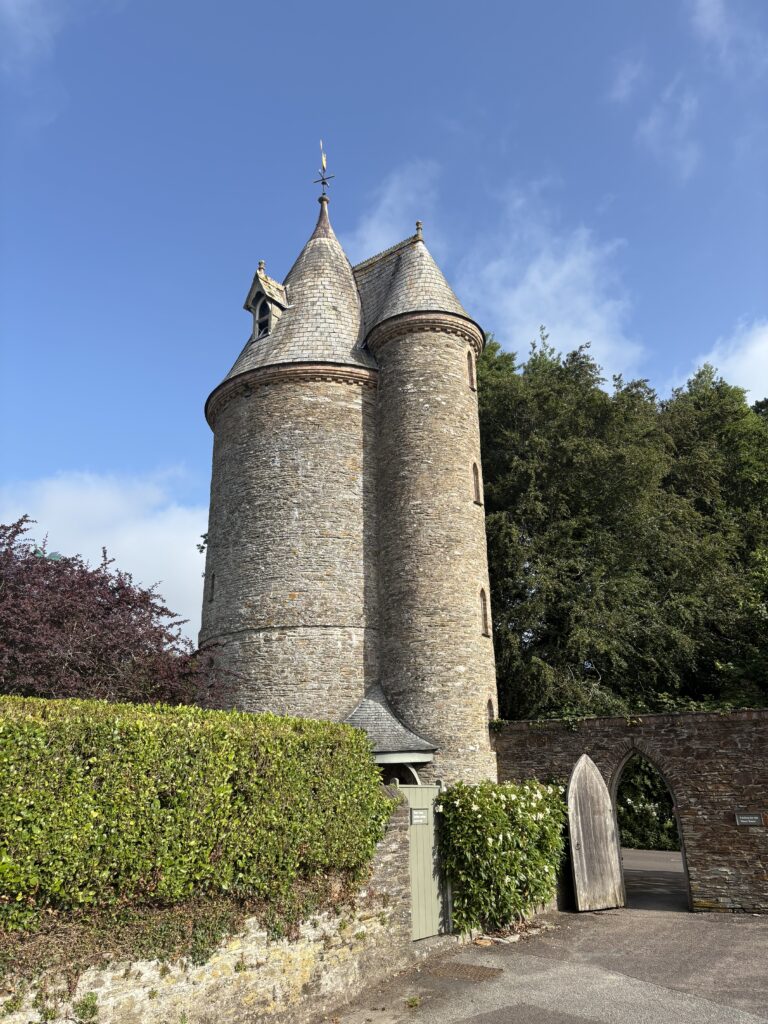 Trelissick parkrun, Cornwall, Parkrun, Parkrun tourism, National Trust Trelissick, A narrow castle-like building stands behind a hedge and in front of trees and a blue sky next to a wall with an open door