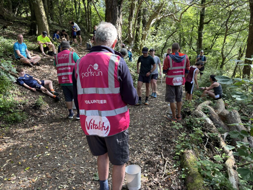 Trelissick parkrun, Parkrun, Parkrun tourism, Runners, A forest with three people wearing pink high-vis jackets seen from behind which say parkrun volunteer and lots of runners sitting down on the ground in the forest