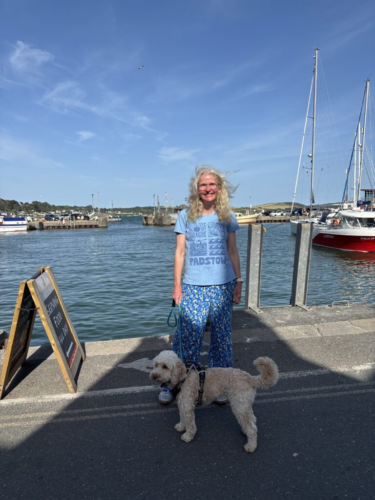 Padstow, Holiday, Hetty, Cockapoo, A woman in a blue Tshirt which says Padstow stands with a golden cockapoo dog in front of the sea and a blue sky with boats in the background