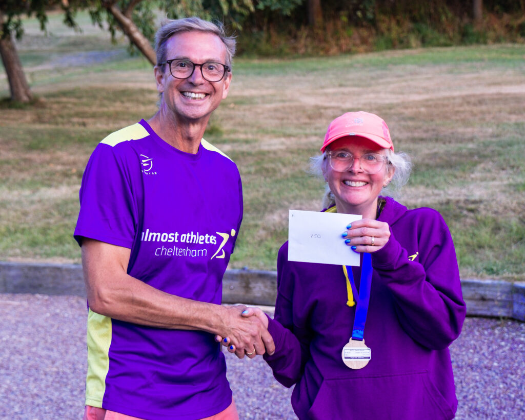 Bugatti 10k prize, Bugatti 10k, Running, Runner, A smiling man in glasses and a purple Tshirt shakes hands with a smiling woman in a purple hoodie and pink cap holding an envelope