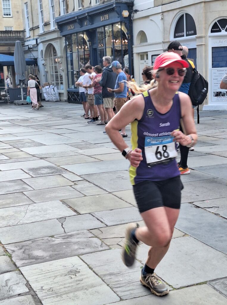 Cotswold Way Relay, Cotswold Way, Bath, Bath city centre, Running, Runner, A female runner in a purple vest pink hat and sunglasses runs through a city centre