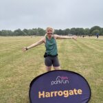 Harrogate parkrun, Parkrun, Parkrun tourism, Running, Runner, A smiling female runner wearing a green vest stands on scorched grass with trees in the distance behind a large purple sign which says parkrun and Harrogate in yellow letters