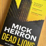 Dead Lions by Mick Herron, Dead Lions, Mick Herron, A bright yellow book called Dead Lions by Mick Herron with the silhouette of a skyscraper on the front sits on a wooden table