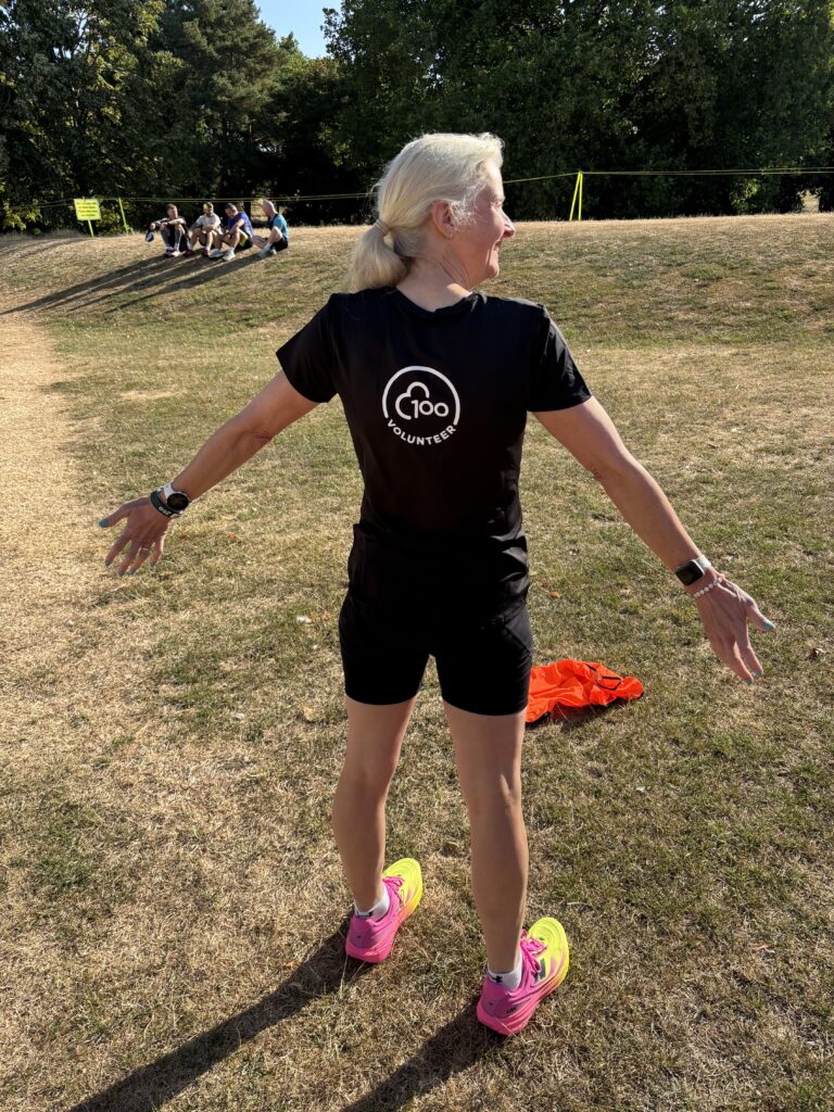 100th volunteer milestone Tshirt, Volunteering, Parkrun, A woman stands with her back to the camera on scorched grass wearing pink and yellow trainers back shorts and a black Tshirt with a white logo on the back saying 100 volunteer