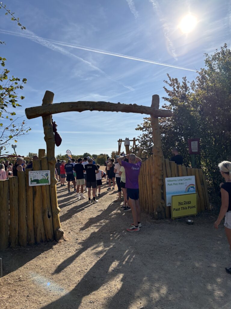 Kilkenny Lane Country parkrun, parkrun, parkrun tourism, A rustic wooden archway with a crowd of people in shorts with their backs to the camera in the distance against a blue sky and a sign on the gate says No dogs past this point