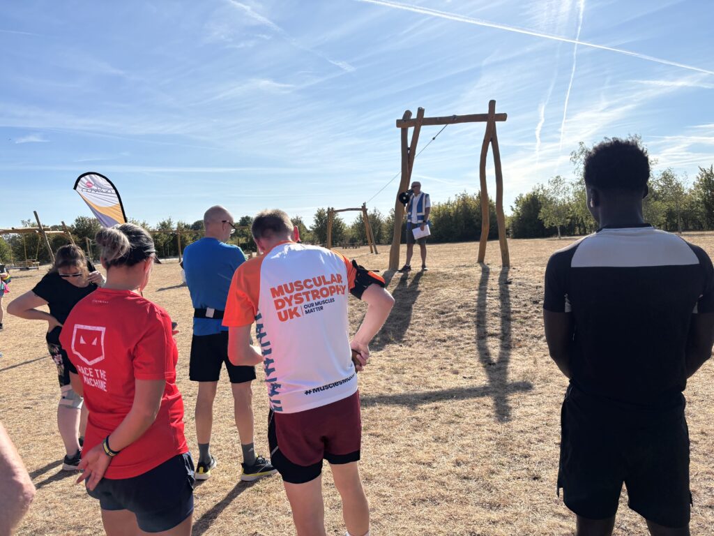 Kilkenny Lane Country parkrun, parkrun tourism, Run director's briefing, A crowd of people stand with their backs to the camera facing a man on raised ground on scorched grass with a loud hailer against a blue sky