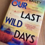 Our Last Wild Days by Anna Bailey, Our Last Wild Days, Anna Bailey, Book review, A book with a multicoloured sunset and a boat amongst reeds on the cover called Our Last Wild Days by Anna Bailey sits on a wooden table