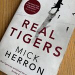 Real Tigers by Mick Herron, Real Tigers, Mick Herron, Book review, A white book called Real Tigers by Mick Herron showing a man and his shadow walking across a paved area surrounded by birds on the cover sits on a wooden table