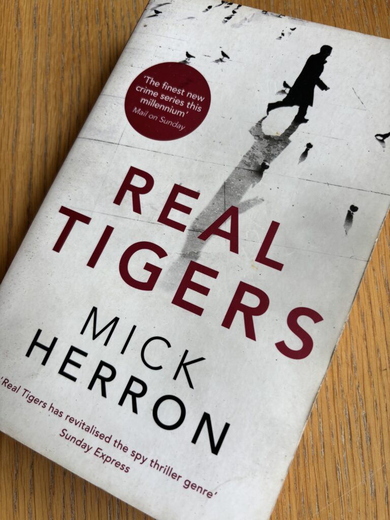 Real Tigers by Mick Herron, Real Tigers, Mick Herron, Book review, A white book called Real Tigers by Mick Herron showing a man and his shadow walking across a paved area surrounded by birds on the cover sits on a wooden table