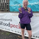 Angels 10k, Running, Runner, A smiling female runner in a purple hoodie and black shorts stands against some big signs holding a medal and a small trophy