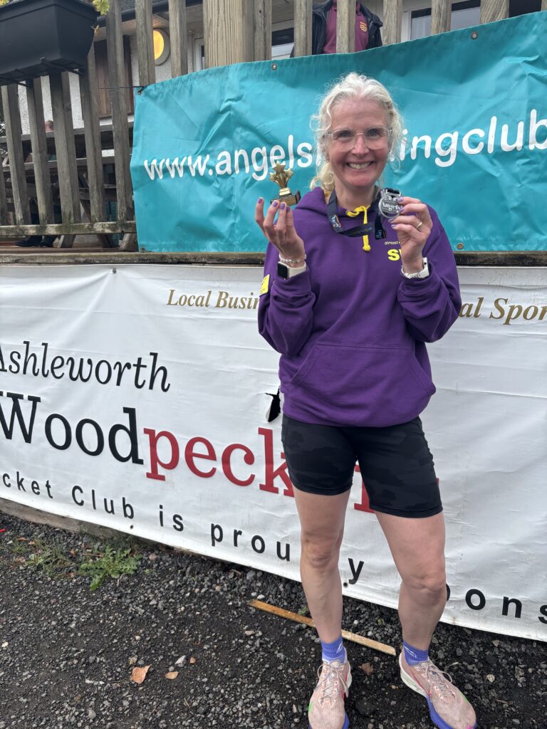 Angels 10k, Running, Runner, A smiling female runner in a purple hoodie and black shorts stands against some big signs holding a medal and a small trophy
