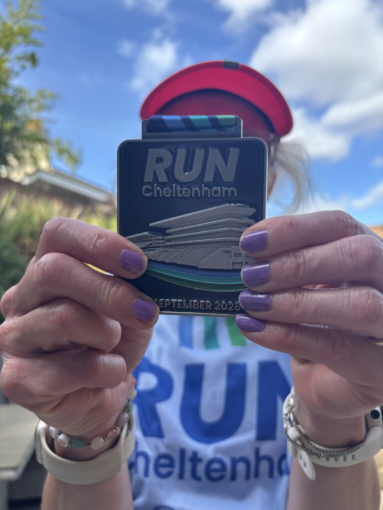 Running, Runner, Cheltenham half marathon, Cheltenham half marathon medal, Close up of a blue metal rectangular medal which says run Cheltenham held by a woman with purple nails and her Run Cheltenham Tshirt just visible in the background