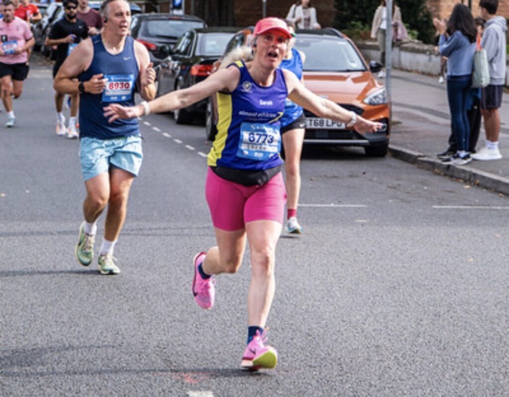Running, Runner, Cheltenham half marathon, A female runner in a purple vest with pink cap and shorts and trainers runs along a road with her arms wide with other runners and parked cars in the background