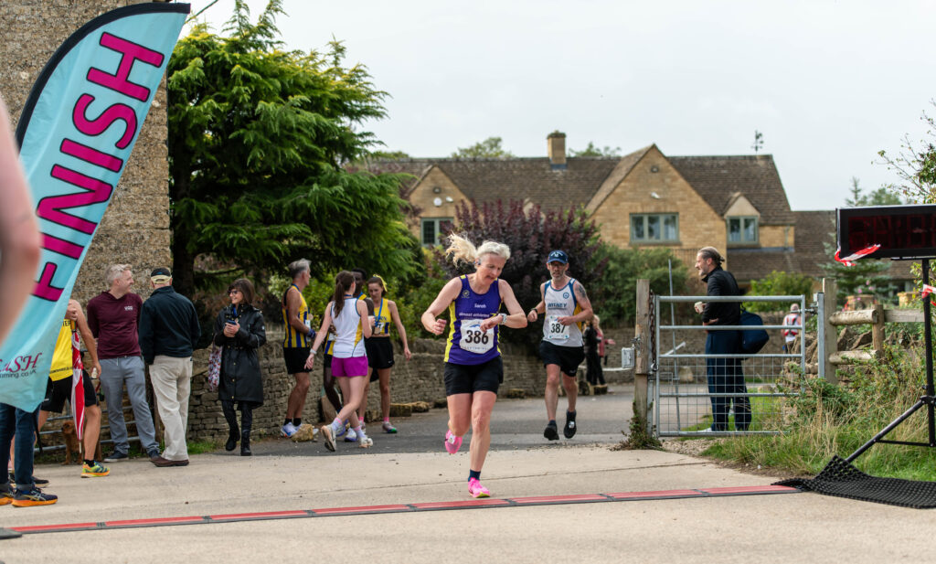 Bourton half marathon, Running, Runner, A female runner in a purple vest crosses a finish line with a male runer and lots of people and a Cotswold stone building in the background