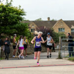 Bourton half marathon, Running, Runner, A female runner in a purple vest crosses a finish line with a male runer and lots of people and a Cotswold stone building in the background