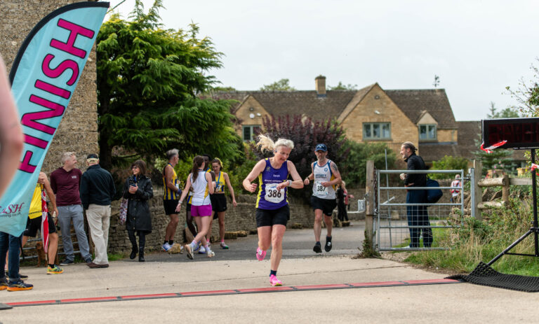 Bourton half marathon, Running, Runner, A female runner in a purple vest crosses a finish line with a male runer and lots of people and a Cotswold stone building in the background