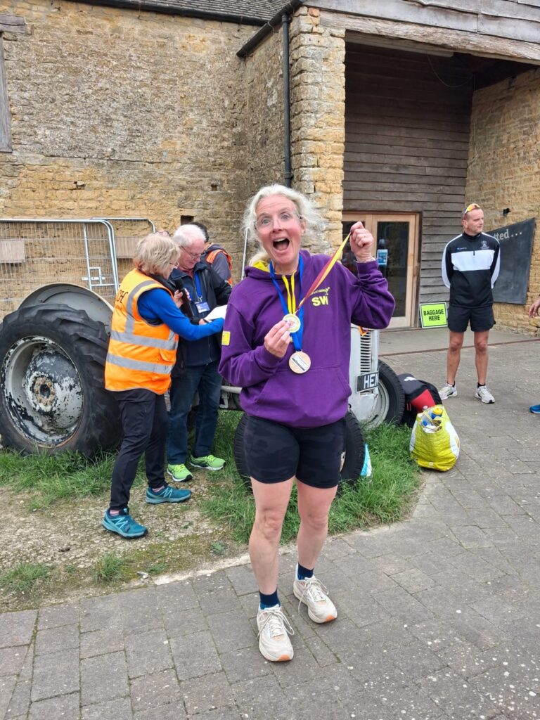 Bourton half marathon, Medals, Running, Runner, A smiling woman in a purple hoodie and black shorts has a medal round her neck and another in her hands against a Cotswold stone wall with runners and officials in the background