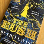 The Rush by Beth Lewis, The Rush, Beth Lewis, Book review, A navy coloured book called The Rush by Beth Lewis with lines of gold across the cover and the silhouette of a woman in a hat and long coat carrying a gun also in gold sits on a wooden table