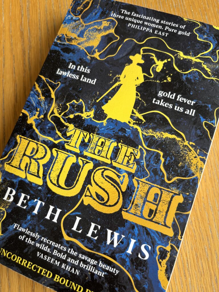 The Rush by Beth Lewis, The Rush, Beth Lewis, Book review, A navy coloured book called The Rush by Beth Lewis with lines of gold across the cover and the silhouette of a woman in a hat and long coat carrying a gun also in gold sits on a wooden table
