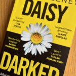 Daisy Darker by Alice Feeney, Daisy Darker, Alice Feeney, Book review, A book called Daisy Darker by Alice Feeney with one half of the cover yellow with black writing and the other half black with yellow writing with a large daisy in the centre sits on a wooden table