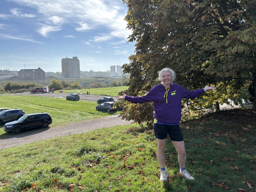 Autumn, Sharpness 4, A smiling woman in a purple hoodie and black shorts stands with her arms wide in front of an autumnal tree with a blue sky and parked cars and tall buildings in the background