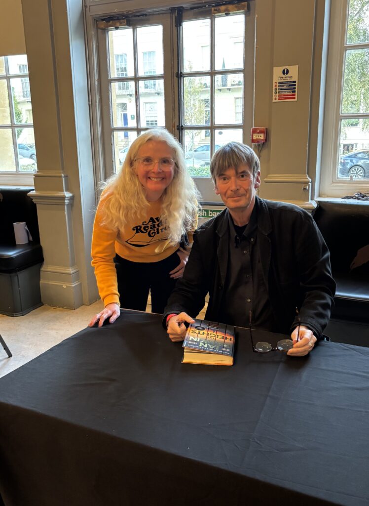 Ian Rankin, Author, Book signing, The Heights by Ian Rankin, A man in his 60s dressed all in black sits behind a black covered table holding a book called The Heights by Ian Rankin and a smiling blonde woman wearing a yellow jumper stands next to him