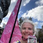 Altra Yorkshire 10 mile race medal, Runner, Medal, A smiling female runner holds up a rectangular medal against a backdrop of a pink a flag and a blue cloudy sky