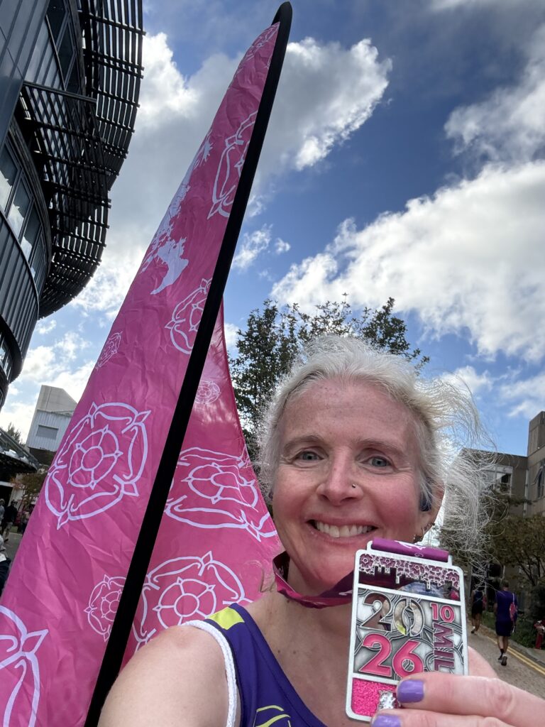 Altra Yorkshire 10 mile race medal, Runner, Medal, A smiling female runner holds up a rectangular medal against a backdrop of a pink a flag and a blue cloudy sky