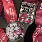Altra Yorkshire marathon medal, Altra Yorkshire 10 mile race medal, Medals, A round silver medal and a rectangular silver medal both with pink detailing and pink ribbons sit on a dark wood surface