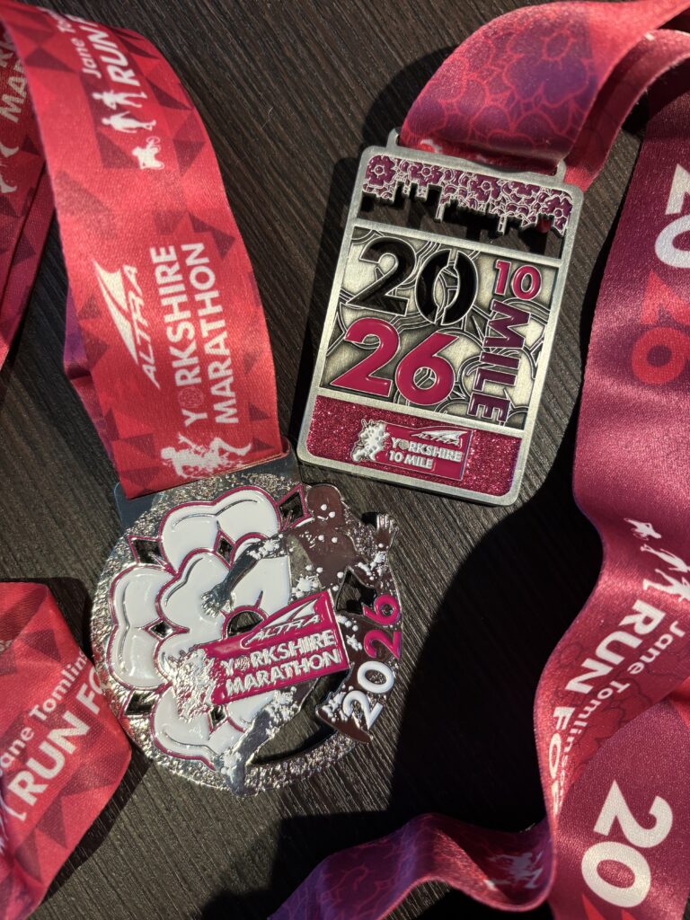 Altra Yorkshire marathon medal, Altra Yorkshire 10 mile race medal, Medals, A round silver medal and a rectangular silver medal both with pink detailing and pink ribbons sit on a dark wood surface